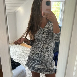 Mönstrad kortklänning med v-ringning - Supersnygg kortklänning med svartvitt mönster och v-ringning. Klänningen har långa ärmar och resår i midjan för en skön passform. Perfekt för dig som gillar en avslappnad men trendig stil.