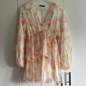 Blommig kortklänning från Gina Tricot - Supersöt kortklänning från Gina Tricot i ljus, transparent polyester med blommigt mönster i rosa, gul och orange. Klänningen har v-ringning, volanger och långa puffärmar. Perfekt för sommarens alla tillfällen!