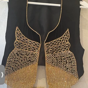 Svart sammetväst med guldpaljetter - Unik svart väst i sammet med detaljerade guldpaljetter och gnistrande pärlor framtill. Västen har öppen front och är dekorerad med glittrande fransar i guld som ger en riktigt festlig vibe. Perfekt för dig som vill sticka ut och addera extra bling till din outfit.