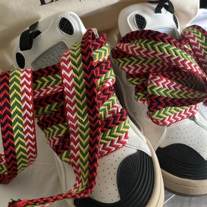 Lanvin sneakers med färgglada snören - Lanvin sneakers i vitt skinn med svarta detaljer och chunky sula. Skorna har unika, breda snören i rött, grönt, gult, svart och vitt med zigzag-mönster. Perforerad tå och broderad bi på sidan ger extra lyxig känsla. Perfekta för dig som vill sticka ut.