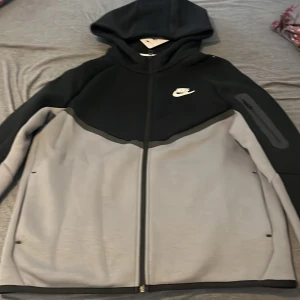 Nike svart och grå hoodiejacka XL - Nike hoodiejacka i svart och grått med dragkedja och huva. Jackan har en clean design med Nike-logga på bröstet och är tillverkad i mjukt syntetmaterial. Perfekt för chill dagar eller när du vill ha en sportig look. Långärmad och med praktiska fickor på sidorna.