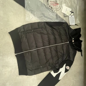 Svart Moncler pufferjacka med mönstrade ärmar -  Helt ny Svart pufferjacka från Moncler med stickade ärmar i svart och vitt zigzag-mönster. Jackan har huva med snörning, hel dragkedja och två fickor framtill. Materialmix av dun, polyester och stickat ger en riktigt schysst look.