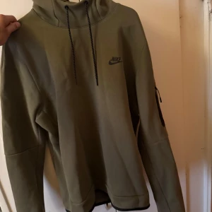 Grön Nike hoodie med huva - Snygg olivgrön hoodie från Nike med svart logga på bröstet. Tröjan har huva med snörning, lång ärm och dragkedjeficka på ena ärmen. Perfekt för chill eller träning, riktigt skön passform och stilren look.