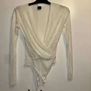 Vit omlott bodysuit från Gina Tricot - Supersnygg vit omlott bodysuit från Gina Tricot i ribbad, glansig polyester. Djup v-ringning framtill och långa ärmar. Perfekt att styla med jeans eller kjol för en trendig look.