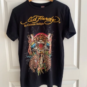 Ed Hardy t-shirt med tryck - Svart t-shirt från Ed Hardy med stort färgglatt tryck på framsidan. Gult Ed Hardy-tryck både fram och bak. Strl L men sitter mer som en stor S eller liten M. Inköpt 2008, inte använd på över 15 år men lagrad i kartong. Klassisk passform och rund hals. Materialet är mjuk bomull. 