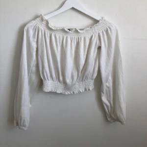Vit offshoulder blus  - Superfin vit blus med offshoulder och långa ärmar. Toppen har smockdetaljer vid halsringning och midja, vilket ger en snygg croppad passform.