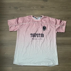 Trapstar t-shirt i rosa och vitt - Säljer en snygg t-shirt från Trapstar i rosa och vitt med coolt tryck framtill. T-shirten har korta ärmar, rund halsringning och en vit piping-detalj över bröstet. Perfekt för dig som gillar streetwear. Storlek M. 