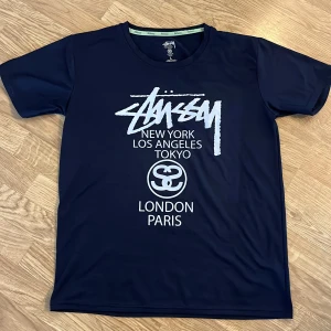 Mörkblå t-shirt från Stüssy - Säljer en mörkblå t-shirt från Stüssy med vit logga och tryck på bröstet med städer som New York, Los Angeles, Tokyo, London och Paris. T-shirten har rund hals och korta ärmar. Perfekt för en streetwear-look.