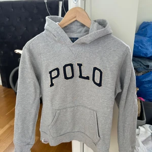 Grå hoodie från Polo Ralph Lauren - Säljer en klassisk grå hoodie från Polo Ralph Lauren med broderad POLO-text på bröstet. Tröjan har huva, magficka och ribbade muddar. Perfekt för en avslappnad och stilren look. (Barn storlek L, 12/14 men passar som dam xs) Inga fläckar, hål eller liknande (obs bara en skugga på bilden dvs ingen fläck)