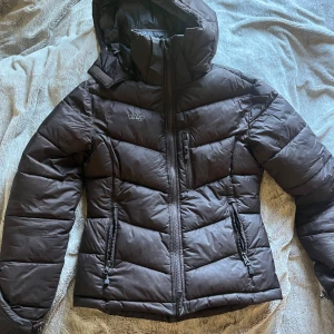 Brun dunjacka från Uhip - Säljer en brun dunjacka från Uhip med huva och dragkedjor framtill samt på fickorna. Jackan har en sportig look och är perfekt för kyliga dagar. Snyggt mönstrat innerfoder och hög krage för extra värme.