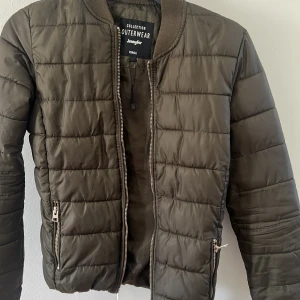 Olivgrön quiltad bomberjacka från Jennyfer -  Går att ta för 100kr.  rSäljer en olivgrön quiltad bomberjacka från Jennyfer i storlek XS. Jackan har dragkedja framtill och vid fickorna samt ribbade muddar vid hals och ärmslut. Perfekt för dig som gillar en stilren och enkel look. 