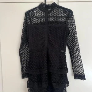 Svart spetsklänning från AX Paris - Säljer en svart kortklänning från AX Paris med långärmade spetsdetaljer och hög hals. Klänningen har flera volanger och dragkedja i ryggen. Perfekt för dig som gillar en elegant och trendig stil. Liten i storlek.