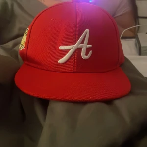 Röd keps med vit A-logga och Los Angeles-märke - Säljer en röd keps med vit broderad A på framsidan och ett Los Angeles Air Sport 1983-märke på sidan. Justerbar snapback baktill. Perfekt för dig som gillar sportig stil.