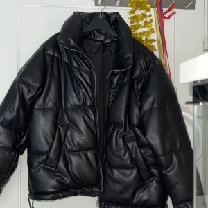 Svart pufferjacka med dragkedja - Säljer en svart pufferjacka med hög krage och dragkedja framtill. Jackan har en oversized passform och är tillverkad i syntetmaterial med quiltad design. Perfekt för kyliga dagar.