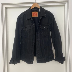 Svart jeansjacka från Levi's - Klassisk svart jeansjacka från Levi's med knappar framtill och två bröstfickor med lock. Jackan har en rak passform och är tillverkad i kraftigt denimtyg. Perfekt för dig som gillar en tidlös och stilren look. Storlek M