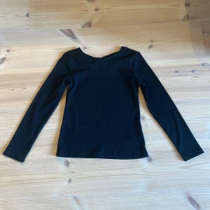 Svart ribbad långärmad topp från Lindex - Säljer en svart ribbad långärmad topp från Lindex med snygg detalj i ryggen. Toppen har en rund halsringning och är tillverkad i ett stretchigt material som sitter skönt på kroppen. Perfekt till många olika outfits!