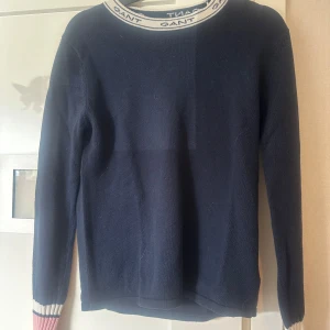 Mörkblå stickad tröja från Gant - Säljer en mörkblå stickad tröja från Gant med vita och röda detaljer vid halsringning och ärmslut. Tröjan har lång ärm och Gant-logga runt halsen. Perfekt för en stilren och klassisk look. Strl 170/s