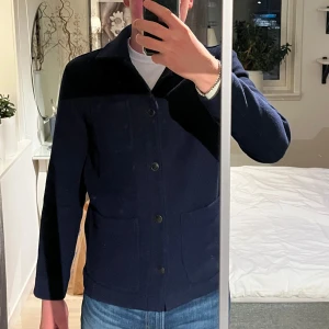 Mörkblå ulljacka från Kappahl - Stilren mörkblå jacka från Kappahl med klassisk krage och tre stora fickor framtill. Jackan har knäppning med knappar och är tillverkad i ullblandning. Perfekt för lager-på-lager och passar till jeans eller chinos. Storlek S men skulle nog säga att den även passar M.