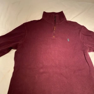Vinröd halvzip tröja från Polo Ralph Lauren - Säljer en vinröd stickad halvzip-tröja från Polo Ralph Lauren med broderad logga på bröstet. Tröjan har hög krage och dragkedja framtill. Perfekt för lager-på-lager och snygg till jeans.