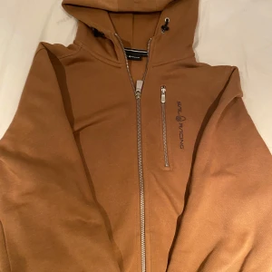 Brun hoodie från Sail Racing - Säljer en brun hoodiejacka från Sail Racing med dragkedja framtill och på bröstfickan. Jackan har huva med snörning och logga på bröstet. Perfekt för dig som gillar sportig stil.