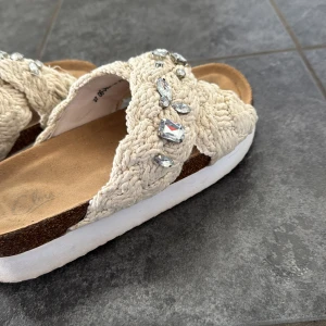Beige sandaler med stickad ovandel och strasstenar - Säljer ett par beige sandaler med stickad ovandel och dekorativa strasstenar. Skorna har en platt vit sula och öppen tå, perfekta för varma dagar. Snygga detaljer som ger en unik look. Använda 3 gånger. 