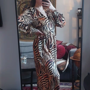 Zebramönstrad långklänning med v-ringning - Säljer en långklänning med zebramönster i svart, beige och orange toner. Klänningen har v-ringning, långa ärmar och markerad midja. Perfekt för dig som vill sticka ut med ett djurmönster och snygg siluett.