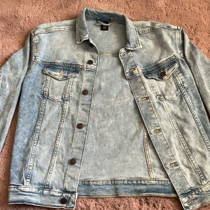 Ljusblå jeansjacka från H&M - Säljer en klassisk ljusblå jeansjacka från H&M. Jackan har knappar framtill, två bröstfickor med lock och knapp samt långa ärmar med knapp vid ärmslut. Perfekt till lager-på-lager och enkel att styla.