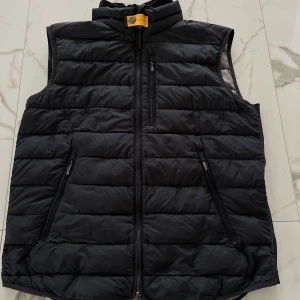 Svart dunväst från Parajumpers - Säljer en svart dunväst från Parajumpers i storlek M. Västen har quiltad design, dragkedja framtill och tre fickor med dragkedja. Lättviktig och perfekt för lager-på-lager. Märket syns tydligt på insidan.