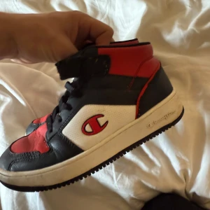 Champion sneakers i svart, rött och vitt - Säljer ett par höga sneakers från Champion i svart, rött och vitt. Skorna har snörning och kardborreband upptill samt rund tå och platt sula. Perfekta för dig som gillar sportig stil.