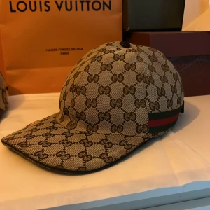 Beige keps från Gucci med monogram - Säljer en beige keps från Gucci med klassiskt GG-monogram över hela kepsen och en grön-röd rand på sidan. Kepsen har böjd skärm och justerbar rem baktill. Perfekt för dig som gillar exklusiva accessoarer.