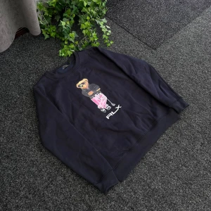 Svart sweatshirt från Polo Ralph Lauren - Cool svart sweatshirt från Polo Ralph Lauren med en ikonisk björn i hoodie och rosa byxor på bröstet. RLX-tryck under motivet. Tillverkad i 100% bomull och har en klassisk passform med rund hals och långa ärmar.