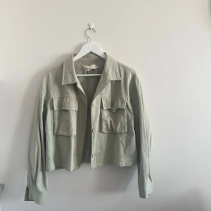 Ljusbeige croppad overshirt med fickor - Säljer en ljusgrön croppad overshirt med klassisk krage och stora bröstfickor med lock. Jackan har knäppning framtill och långa ärmar. Perfekt för lager-på-lager och enkel att styla till olika outfits.