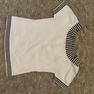 Vit topp med svartvita ränder från MiyaQ - Säljer en vit topp från MiyaQ med svartvita ränder på krage, ärmslut och nederkant. Toppen har korta ärmar och en liten glittrig detalj framtill. Perfekt för en stilren och sportig look.