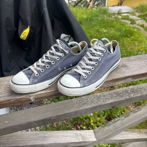 Mörkblåa Converse - Säljer ett par klassiska grå Converse All Star sneakers med vita gummidetaljer och snörning. Skorna har låg modell och rund tå, perfekt för en avslappnad stil. Converse-logga syns baktill på sulan. Helt nya!!