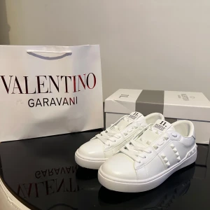 Vita sneakers från Valentino Garavani - Säljer ett par fräscha vita sneakers från Valentino Garavani med ikoniska nitar längs sidorna och bak. Skorna har rund tå, platt sula och är tillverkade i skinn. Perfekta för dig som vill ha en clean och lyxig look.