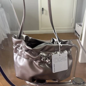 axelväska från C.W. - Säljer en snygg axelväska i metallic silver från C.W. Väskan har dragsko upptill med metallringar och pärldetaljer. Perfekt storlek för det viktigaste och har en trendig, glansig finish. Supersnygg accessoar till din outfit! Helt ny och säljer då den inte kommer till användning. Pris går att diskutera.❤️