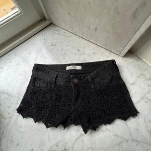 Snygga hollister shorts - Svarta jeansshorts från Hollister med virkad spetsdetalj framtill och råa kanter. Klassisk femficksmodell med knapp och dragkedja. Perfekta för sommaren och ger en cool, bohemisk vibe.