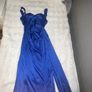 Blå långklänning med strassdetaljer - Säljer en elegant blå långklänning i glansigt satinliknande material. Klänningen har breda axelband, markerad byst och vackra strassdetaljer längs överdelen. Perfekt för bal eller festliga tillfällen.