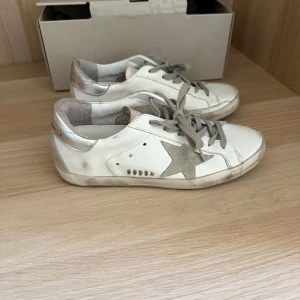 Golden Goose sneakers - Säljer ett par vita sneakers från Golden Goose med grå snörning och grå stjärna på sidan. Skorna har silvriga detaljer vid hälen och klassisk låg modell. Perfekta för dig som gillar en stilren och trendig look. Lite sliten i hälen (privat för bilder, men inget som påverkar bekvämligheten eller utseendet, använda fåtalet gånger)