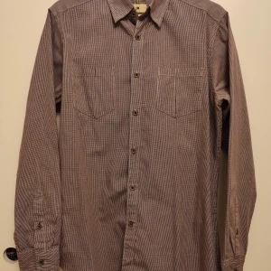 Rutig skjorta från Life Khaki - Säljer en stilren rutig skjorta från Life Khaki i slim fit. Skjortan har lång ärm, knappar framtill och två bröstfickor. Färgen är en mix av brunt, vitt och rött, vilket ger ett klassiskt och tidlöst intryck.