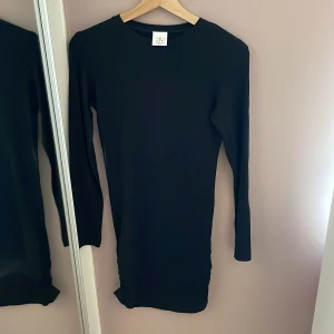 Svart långärmad klänning med rynk från THE NEW - Svart långärmad klänning från THE NEW med tight passform och snygga rynkade detaljer längst ner på sida . Enkel och stilren design som passar perfekt till många tillfällen. Mjuk och stretchig känsla.
