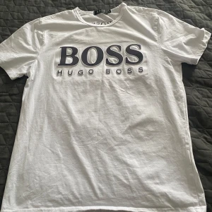 Vit t-shirt från Hugo Boss - Säljer en klassisk vit t-shirt från Hugo Boss med stor svart logga framtill och mindre logga bak i nacken. T-shirten har normal passform och är tillverkad i mjuk bomull. Perfekt för en stilren och enkel look. Behöver bara strykas lite. 