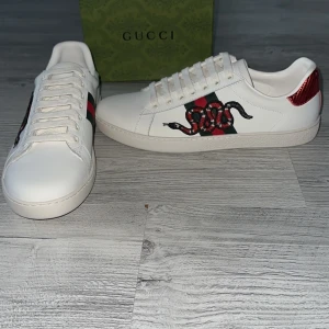 Gucci Ace sneakers med orm - Säljer ett par vita Gucci Ace sneakers med klassiska gröna och röda ränder samt en broderad orm på sidan. Baksidan har detaljer i rött och grönt med Gucci-logga. Snygg och ikonisk modell med rund tå och platt sula