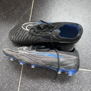 Nike Phantom gx Elite - Säljer ett par Nike Phantom gx i svart med blå detaljer och mönstrad ovandel. Skorna har snörning och en platt sula med fasta dobbar för spel på gräs. Perfekta för dig som vill ha bra grepp och kontroll på planen. Se bilder för defekter, hör av dig vid minsta fundering och skäms inte!🔥💥💯