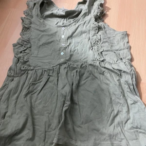 Grön ärmlös blus med spetsdetaljer från Beloved Woman - Säljer en ljusgrön ärmlös blus från Beloved Woman med fina spetsdetaljer längs axlarna och knappar framtill. Blusen har en lös passform och volangdetaljer som ger en romantisk känsla.