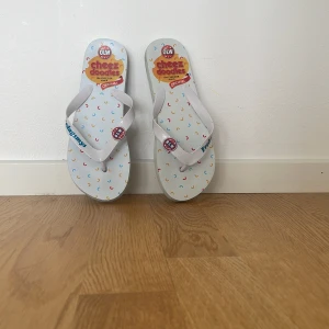 Vita flip-flops med Cheez Doodles-motiv - Unika vita flip-flops med färgglatt Cheez Doodles-mönster och text på remmarna. Perfekta för sommaren och strandhäng. Lätta och bekväma med platt sula och öppen tå.