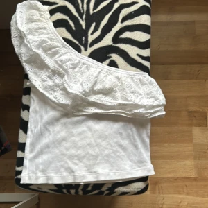 Vit one shoulder topp med volang från Gina Tricot - Säljer en vit one shoulder topp från Gina Tricot i ribbad bomull med bred volang i broderad spets. Perfekt för dig som gillar en trendig och söt stil. Toppen är ärmlös och har en snygg omlottkänsla med volangdetalj. Tröjan har fått en liten solkräms fläck på ena sidan men det syns knappt på. Använd få antal gånger. Perfekt i sommar! 