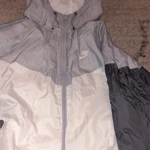 Nike vindjacka i grått och vitt - Säljer en snygg Nike vindjacka i två nyanser av grått och vitt. Jackan har huva med dragsko, dragkedja framtill och Nike-logga på bröstet. Perfekt för dig som gillar sportig stil och vill ha något lätt och smidigt.