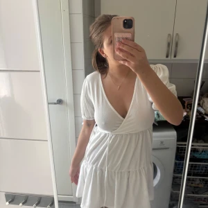 Vit kortklänning med v-ringning från Gina Tricot - Supersöt vit kortklänning från Gina Tricot med djup v-ringning och puffärmar. Klänningen har en volang nedtill och är tillverkad i ett lätt, strukturerat material. Perfekt för sommarens alla tillfällen!