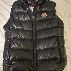 Svart dunväst från Moncler - Säljer en svart dunväst från Moncler med dragkedja och hög krage. Västen är quiltad för extra värme. Perfekt för lager-på-lager och stilren look.